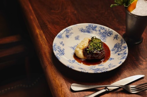 A plate of meat with green herb sauce on a bed of creamy mashed potatoes, set on a wooden table.