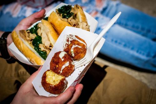 A person holds two sandwiches filled with greens and meat, alongside a container of golden, crispy croquettes.