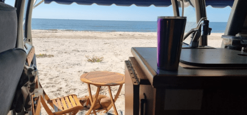 A beach view from a van, featuring a table and chair on the sand, with a colorful cup on the counter inside.