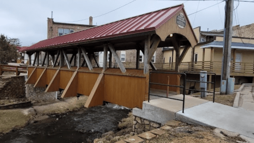 A wooden covered bridge with a red roof spans a small stream, surrounded by buildings and a pathway.