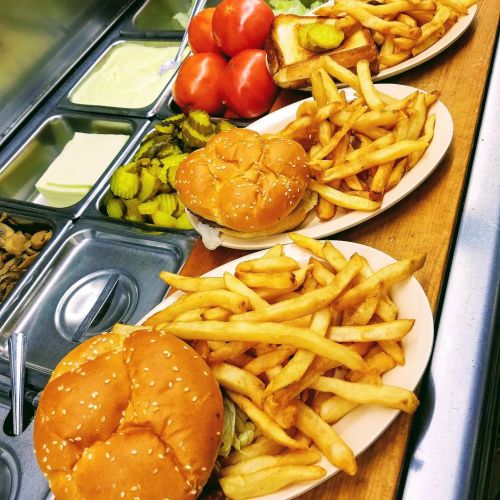 Three plates of burgers with fries, accompanied by tomatoes, pickles, and a slice of toast on a wooden counter.
