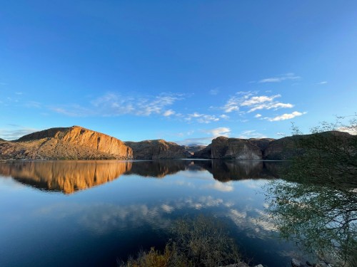 A serene lake reflects rocky cliffs and a clear blue sky, with scattered clouds and greenery in the foreground.