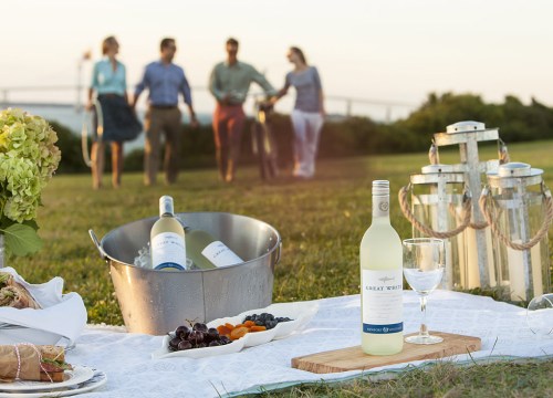 A picnic scene with wine, food, and lanterns, as friends walk in the background during sunset.