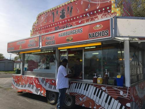 A colorful food truck with a menu featuring hamburgers, nachos, and Philly cheese, with a person ordering at the counter.