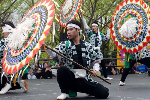 A performer in traditional attire kneels with a stick, surrounded by others holding colorful, decorative fans during a dance.