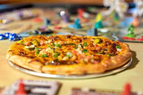 A close-up of a pizza topped with vegetables, placed on a table with colorful game pieces and a board game in the background.