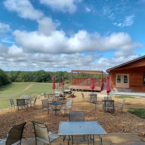 A scenic outdoor area with tables, chairs, and umbrellas, featuring a wooden building and a grassy landscape in the background.