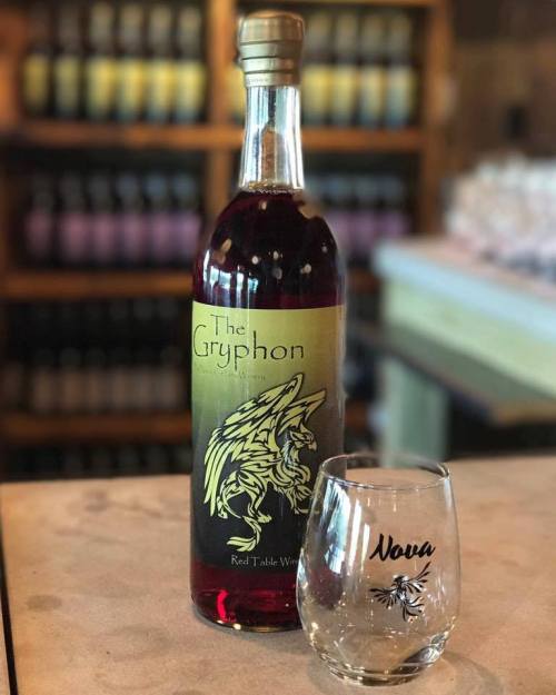 A bottle of "The Gryphon" red table wine next to a glass with "Nova" printed on it, set against a blurred background.