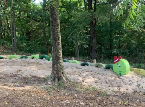 A playful green caterpillar with a red hat is nestled in a sandy area surrounded by trees.