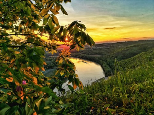 A vibrant sunset over a river, framed by lush green leaves and rolling hills in the background.