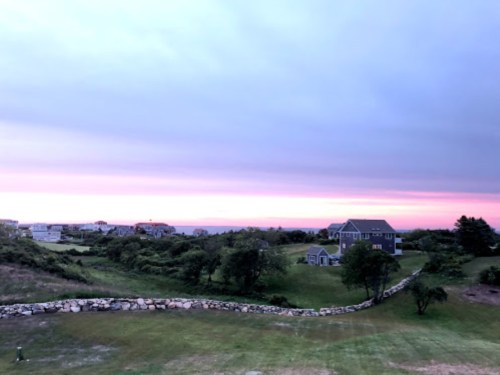 A serene landscape at dusk, featuring a grassy area, stone wall, and houses under a colorful sky.