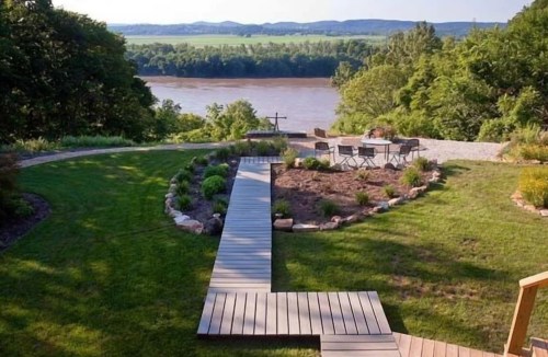 A scenic view of a landscaped garden with a wooden path leading to a river and seating area surrounded by greenery.