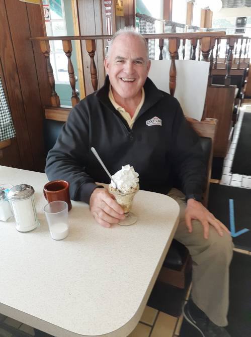 A smiling man sits at a table in a diner, holding a sundae topped with whipped cream.