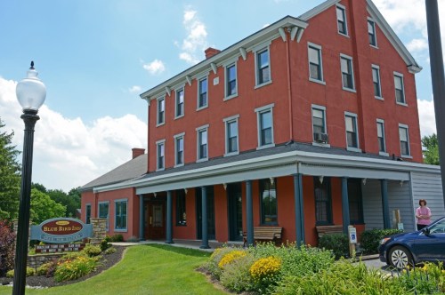 A red brick building with a porch, surrounded by greenery and flowers, under a blue sky with fluffy clouds.