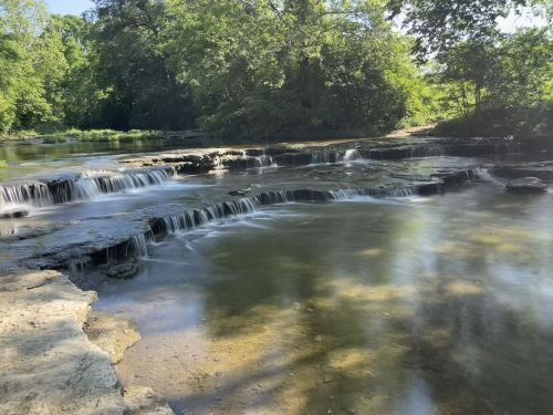 A serene river scene with gentle waterfalls cascading over rocky ledges, surrounded by lush greenery and clear water.