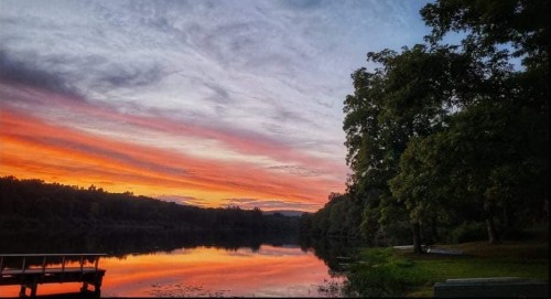 A serene lake at sunset, reflecting vibrant orange and purple skies, surrounded by lush trees.