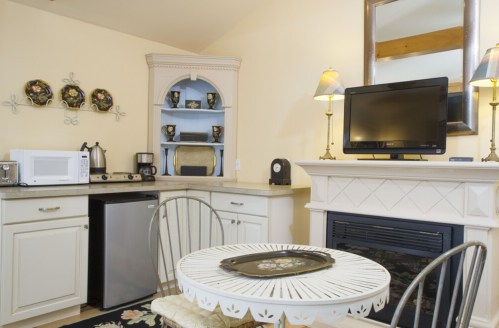 Cozy kitchen area with a small dining table, microwave, fridge, and a TV above a fireplace. Decorated in soft colors.