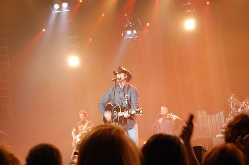 A musician in a cowboy hat performs on stage with a guitar, accompanied by band members and bright stage lights.