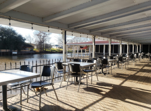 A riverside dining area with empty tables and chairs, featuring sunlight and shadows on the wooden floor.