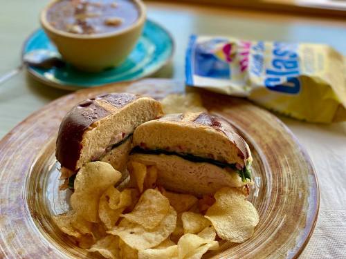 A plate with a cut sandwich, potato chips, and a bowl of soup, with a bag of chips in the background.
