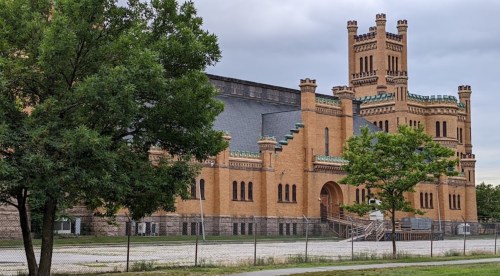 Historic brick building with towers, surrounded by trees and a fence, under a cloudy sky.