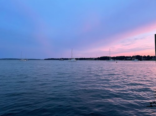 A serene waterfront scene at dusk, featuring sailboats on calm water under a colorful sky.