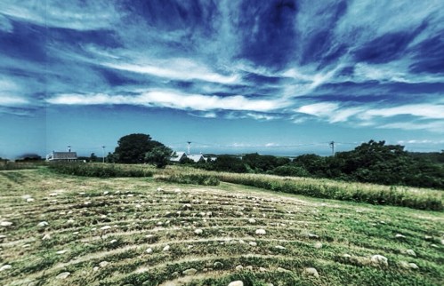 A grassy landscape with stone circles under a dramatic sky filled with clouds. Trees and distant structures are visible.