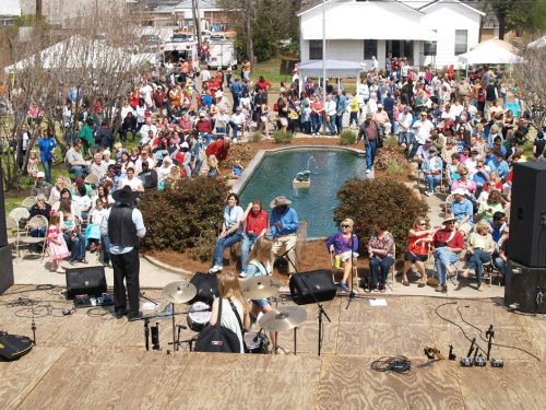 A large crowd gathers around a stage with musicians performing, near a pond in a park setting.