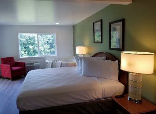 A cozy hotel room featuring a bed, two lamps, a red chair, and a large window with a view of greenery outside.