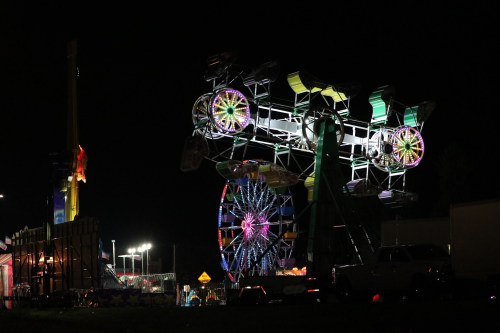 A brightly lit carnival at night featuring a Ferris wheel and a spinning ride against a dark sky.
