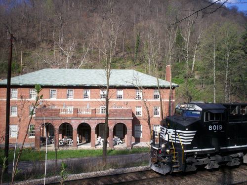 A train passes by a brick building surrounded by trees, with outdoor seating visible on a sunny day.