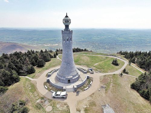 Aerial view of a tall stone tower on a grassy hill, surrounded by trees and vehicles, with a scenic landscape in the background.