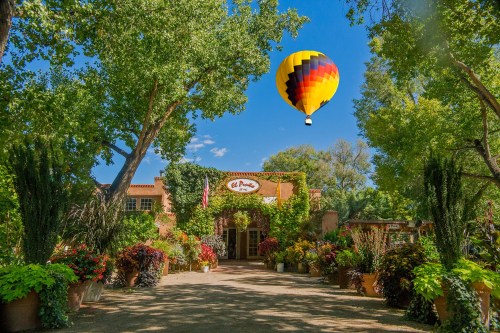 A colorful hot air balloon floats above a lush garden entrance with vibrant flowers and greenery.