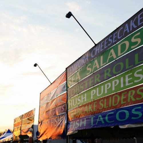 Colorful food vendor signs at sunset, featuring items like tempura cheesecake, lobster rolls, and fish tacos.