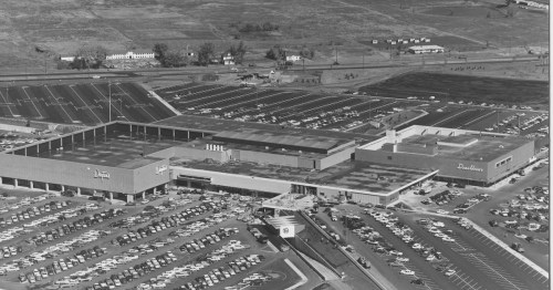 Aerial view of a large shopping mall with extensive parking and surrounding landscape in black and white.