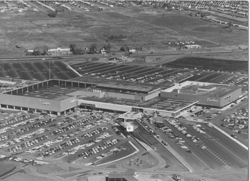 Aerial view of a large shopping center with extensive parking and surrounding landscape in black and white.