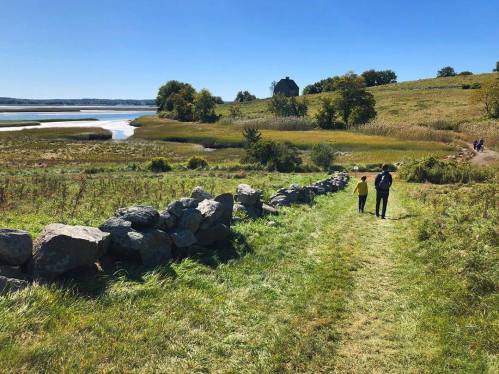 Two people walk along a grassy path beside a stone wall, with a scenic view of fields and water in the background.