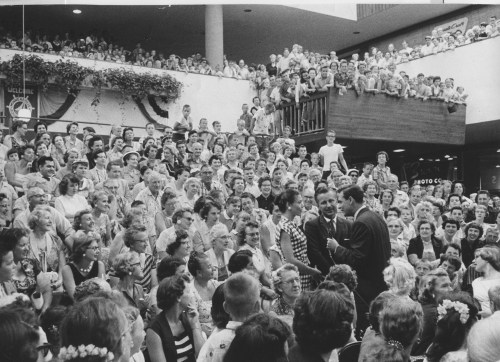 A large crowd gathers in a public space, watching two men speaking on stage, surrounded by decorations and banners.