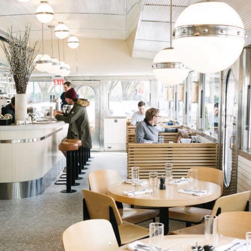 A modern diner interior with round lights, wooden seating, and patrons at the counter and tables.