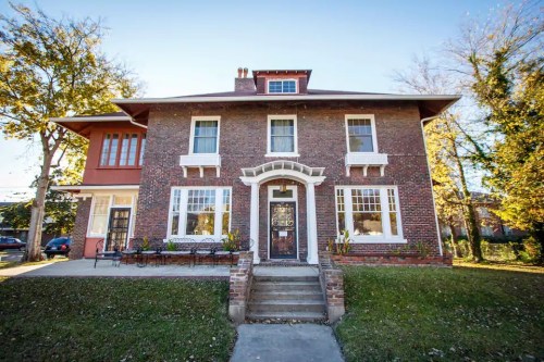 A two-story brick house with large windows, a front porch, and a well-maintained lawn surrounded by trees.