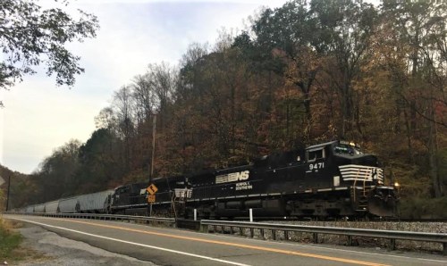 A black train with freight cars travels along a road, surrounded by autumn-colored trees and hills.
