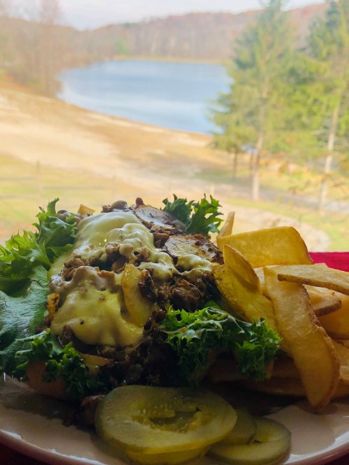 A hearty sandwich with lettuce, cheese, and pickles, served with fries, against a scenic lakeside backdrop.