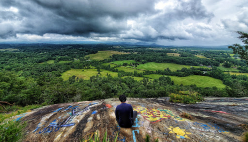 A person sits on a rocky ledge, overlooking a lush green landscape under a cloudy sky.