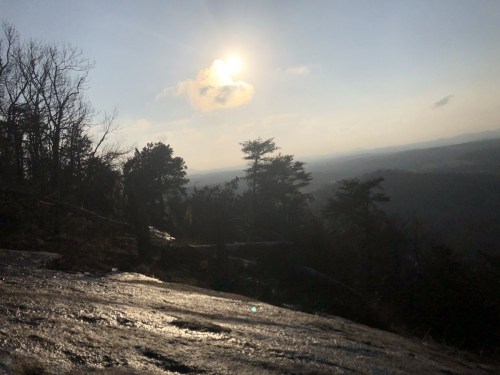 A sunlit landscape with a rocky foreground, trees, and distant mountains under a clear sky.
