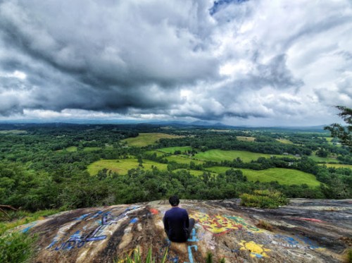 A person sits on a rocky outcrop, overlooking a lush green landscape under a dramatic, cloudy sky.