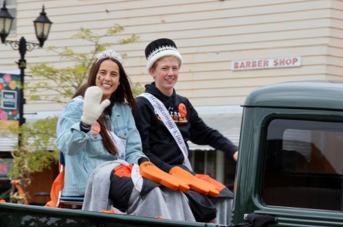 Two crowned individuals wave from the back of a truck during a parade, wearing sashes and smiling at the camera.