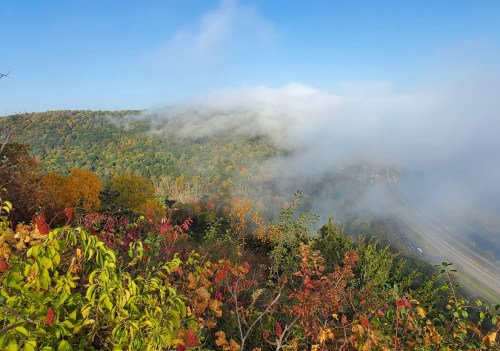A scenic view of a colorful autumn landscape with fog rolling over the hills under a clear blue sky.