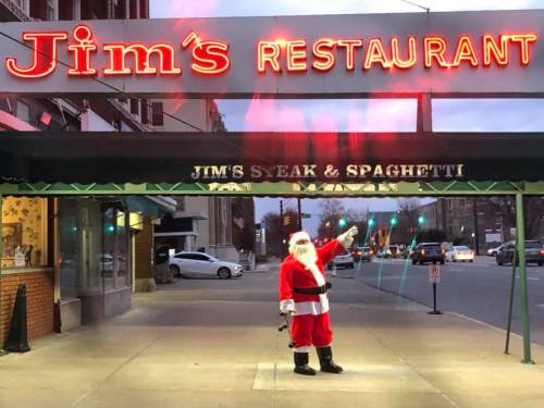 A man dressed as Santa stands outside Jim's Restaurant, with a festive sign overhead in the evening light.