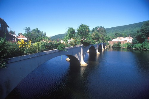 A scenic view of a bridge over a calm river, surrounded by greenery and charming houses under a clear blue sky.