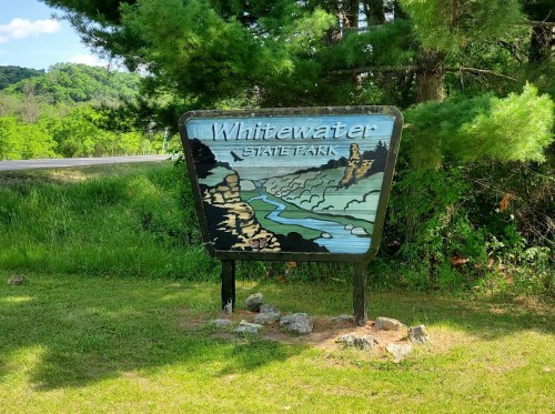 Sign for Whitewater State Park featuring a scenic design with a river and rocky cliffs, surrounded by greenery.